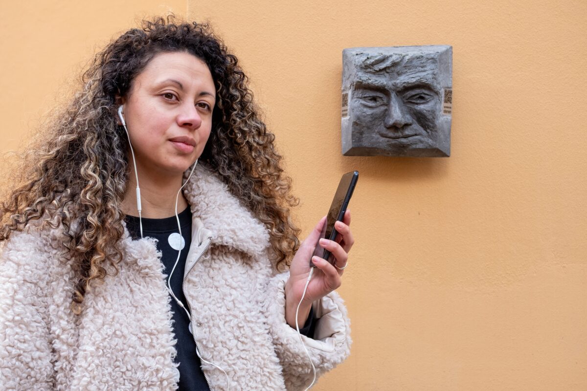 Een vrouw met lang krullend haar, staat naast een Sprekende Steen met haar telefoon in de hand en oordopjes in. Zij draagt een wollig wit vest.