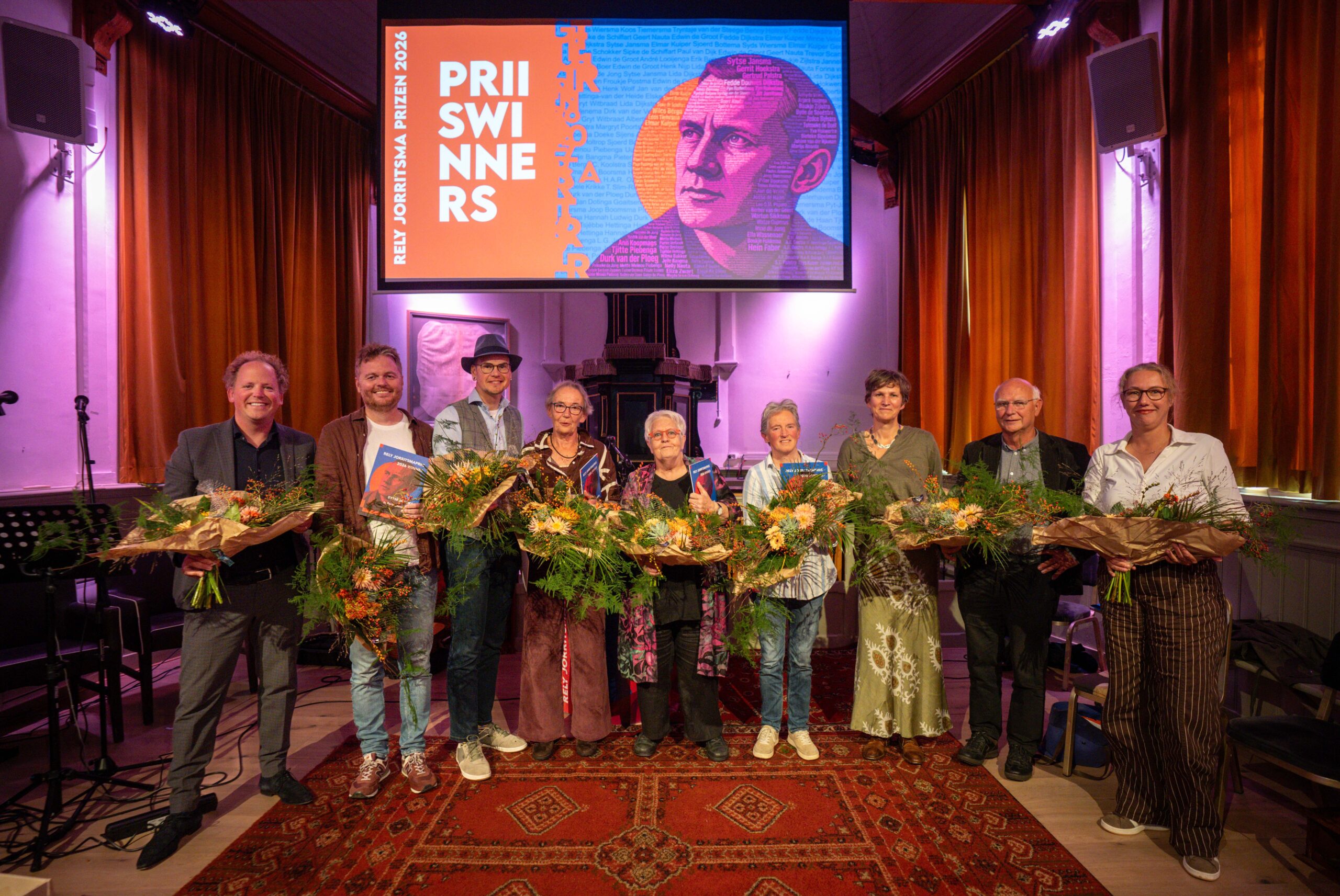 De prijswinnaars en juryleden poseren met bloemen voor een scherm met daarop een foto van Rely Jorritsma