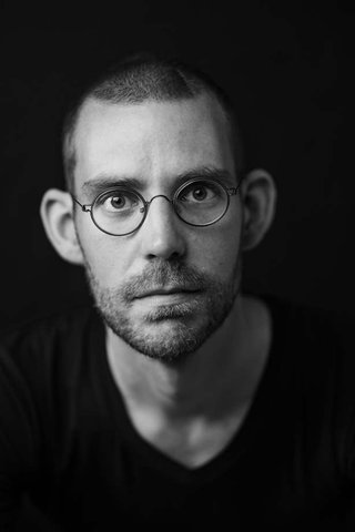 Zwart wit foto van de auteur. Hij heeft kort haar en een jonge baard, een bril met dun montuur, en draagt een zwart shirt.